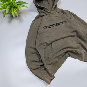 Puede incluir: Una sudadera con capucha verde oliva con capucha y bolsillo delantero. La palabra "carhartt" est&aacute; impresa en negro en el pecho. La sudadera con capucha est&aacute; hecha de un material suave y c&oacute;modo.