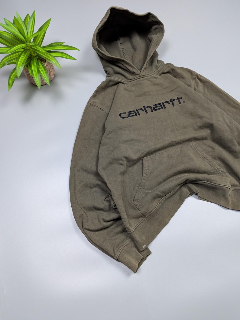 Puede incluir: Una sudadera con capucha verde oliva con capucha y bolsillo delantero. La palabra "carhartt" est&aacute; impresa en negro en el pecho. La sudadera con capucha est&aacute; hecha de un material suave y c&oacute;modo.