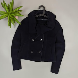 May include: A navy blue double-breasted jacket with a unique, oversized collar. The jacket features two rows of buttons and a tailored fit. The garment is displayed on a black hanger, showcasing its design and structure. A potted plant is visible in the background.