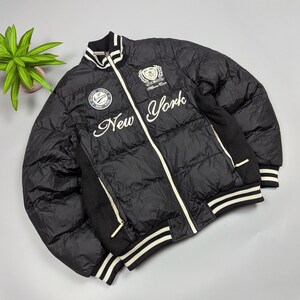 Puede incluir: Chaqueta bomber acolchada negra con detalles blancos y la palabra "New York" bordada en la parte delantera. Cuenta con un panel de punto acanalado en los laterales y las mangas, y ribete a rayas en el cuello, los puños y el dobladillo.