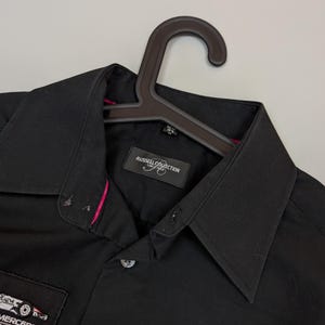 Puede incluir: Camisa negra con cuello y mangas cortas. Tiene parches con texto y logotipos, incluyendo "MERCEDES PARTNER" y "DIAG". La camisa est&aacute; colgada en una percha marr&oacute;n.