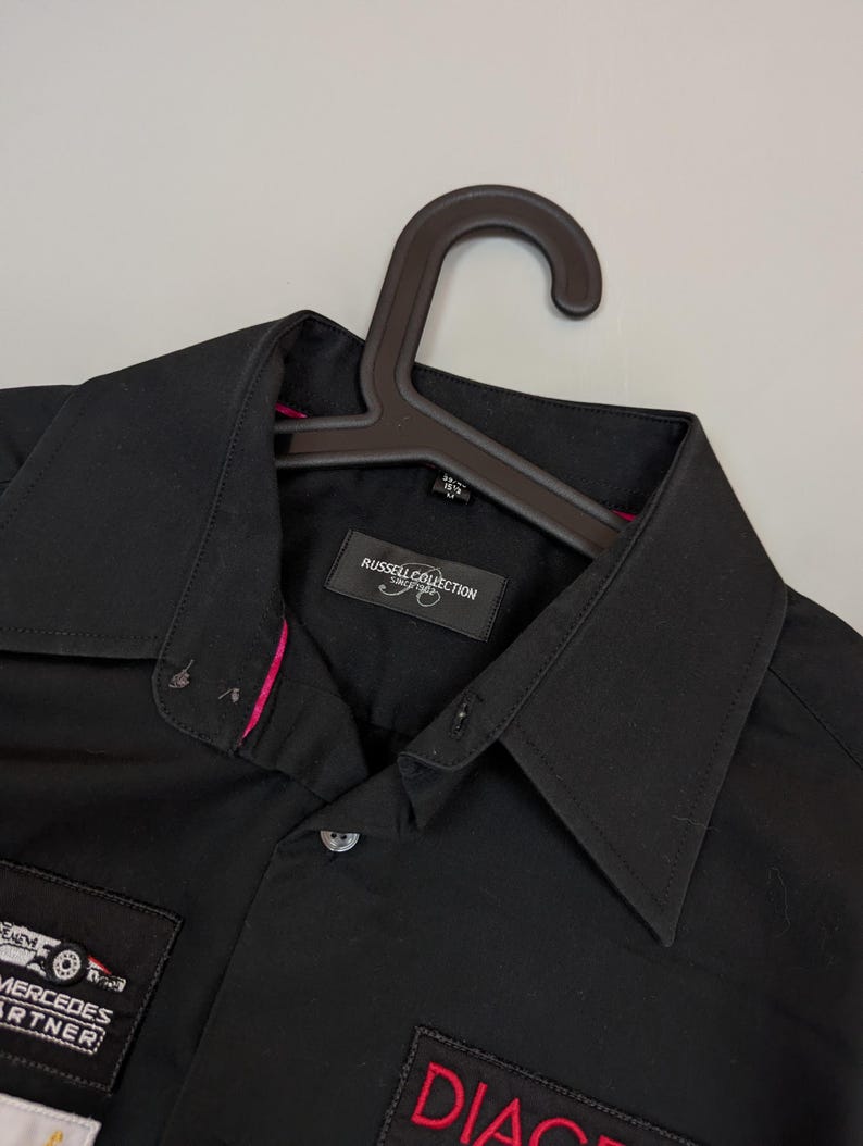 Puede incluir: Camisa negra con cuello y mangas cortas. Tiene parches con texto y logotipos, incluyendo "MERCEDES PARTNER" y "DIAG". La camisa est&aacute; colgada en una percha marr&oacute;n.