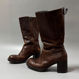 May include: A pair of mid-calf brown leather boots featuring a block heel, a side zipper closure, and a small embossed crest logo on the outer side of the shaft. The boots have a rounded toe and visible stitching details along the leather panels.