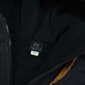 Pode incluir: Um casaco preto com capuz e fecho de correr, forro interior escuro e uma faixa castanha ao longo do fecho. Uma etiqueta preta com um elefante branco e o texto Nomads est&aacute; cosida no interior, com uma etiqueta branca indicando 100% Cotton, Made in Nepal, tamanho L.