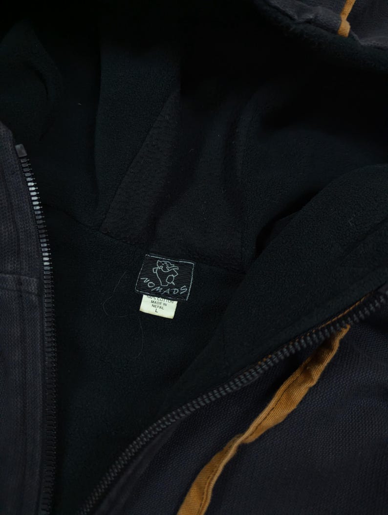 Pode incluir: Um casaco preto com capuz e fecho de correr, forro interior escuro e uma faixa castanha ao longo do fecho. Uma etiqueta preta com um elefante branco e o texto Nomads est&aacute; cosida no interior, com uma etiqueta branca indicando 100% Cotton, Made in Nepal, tamanho L.
