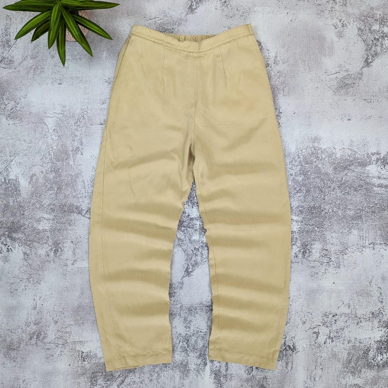 May include: A pair of solid light tan trousers featuring a high-waisted design with an elasticized waistband, a straight-leg cut, and subtle vertical seam detailing running down the front of each leg, presented in a clean and simple style.