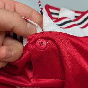 May include: Close-up of a red athletic jersey with a white collar and red button. The collar has black and white stripes. The Adidas logo is visible on the button and the jersey.