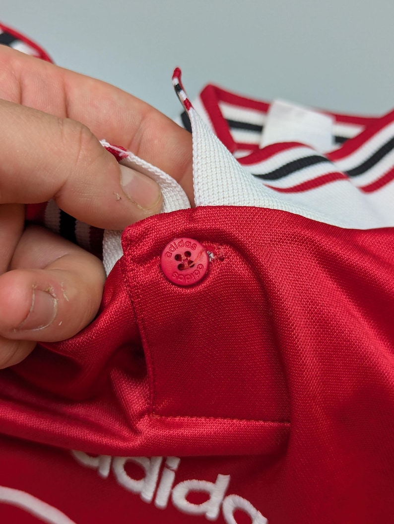 May include: Close-up of a red athletic jersey with a white collar and red button. The collar has black and white stripes. The Adidas logo is visible on the button and the jersey.