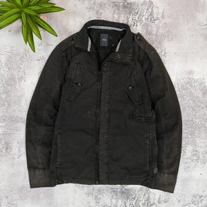 May include: A dark brown jacket with a full-length zipper, multiple pockets, and button details. The jacket has a stand-up collar and a subtle, textured appearance. The overall style is casual and utilitarian.