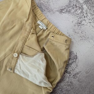 May include: Beige trousers featuring a high-waisted design with an elastic waistband, a front button closure, and a metal hook-and-eye fastener. The interior lining is white, and the garment label reads Michael Glasse. The fabric has a fine texture.