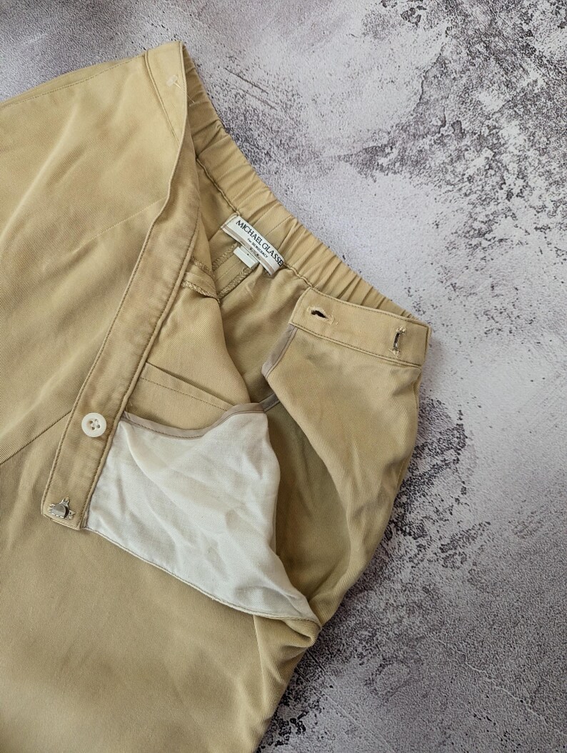 May include: Beige trousers featuring a high-waisted design with an elastic waistband, a front button closure, and a metal hook-and-eye fastener. The interior lining is white, and the garment label reads Michael Glasse. The fabric has a fine texture.