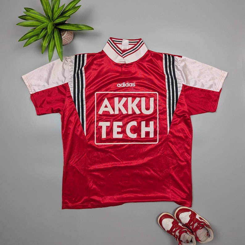 May include: Red Adidas soccer jersey with white sleeves and collar. The jersey features black stripes on the sides and the text "AKKU TECH" in a white box on the front. A pair of red and white sneakers are at the bottom.