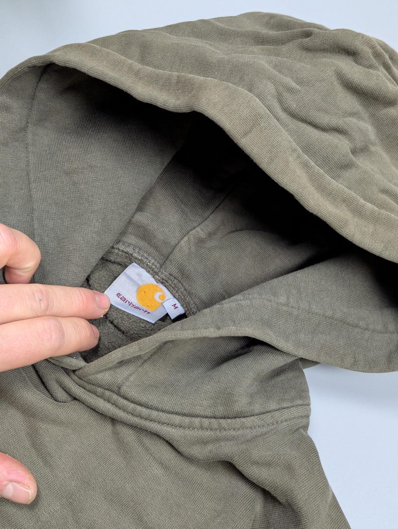 Puede incluir: Sudadera con capucha verde oliva con cord&oacute;n. El logotipo de Carhartt es visible en una etiqueta dentro de la capucha. La sudadera est&aacute; hecha de un material suave y c&oacute;modo.