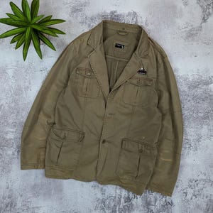 May include: A khaki-colored jacket with multiple front pockets and a button closure. The jacket has a collared neckline and a small emblem on the chest pocket. The jacket is made of a durable fabric and has a tailored fit.