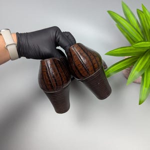 Może przedstawiać: Para brązowych but&oacute;w na obcasie z teksturowanym wzorem przypominającym krokodyla. Buty mają zaokrąglony nosek i gruby obcas. Buty są trzymane przez dłoń w rękawicy, z rośliną w tle.