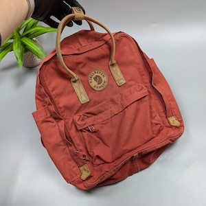 May include: A rust-colored backpack with a front pocket and side pocket. The backpack has a top handle and leather accents. The brand name "Fjallraven Kanken" is visible on a circular patch on the front of the bag.