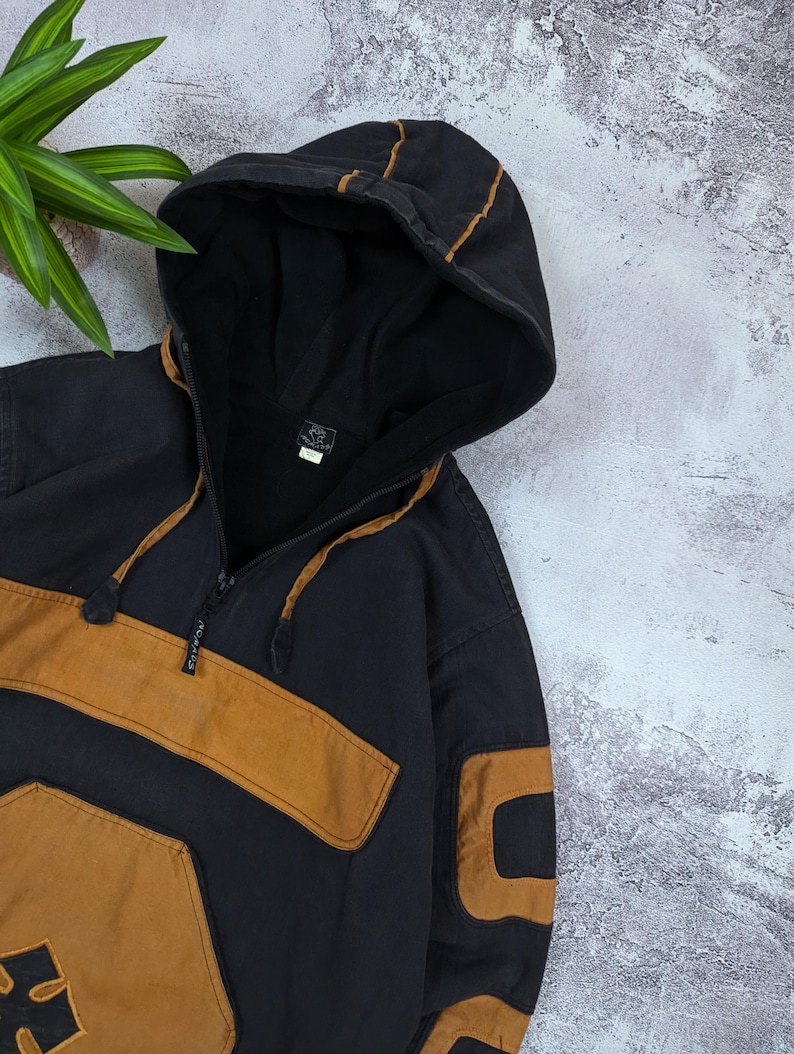 Pode incluir: Um moletom com capuz preto e castanho com fecho de meio z&iacute;per no pesco&ccedil;o, um bolso frontal grande e mangas compridas com pain&eacute;is castanhos. A pe&ccedil;a inclui um capuz com cord&atilde;o e uma pequena etiqueta da marca no interior da gola.