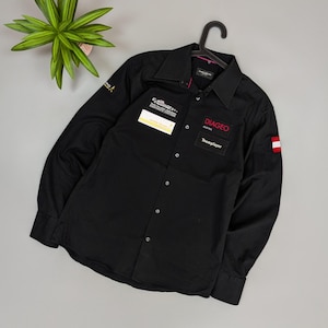 Puede incluir: Camisa negra de manga larga con cuello. Presenta parches con texto y logotipos, incluyendo "DIAGEO AUSTRIA Teamplayer" en rojo. Un parche con la bandera austriaca est&aacute; en la manga derecha. La camisa est&aacute; colgada de una percha negra.