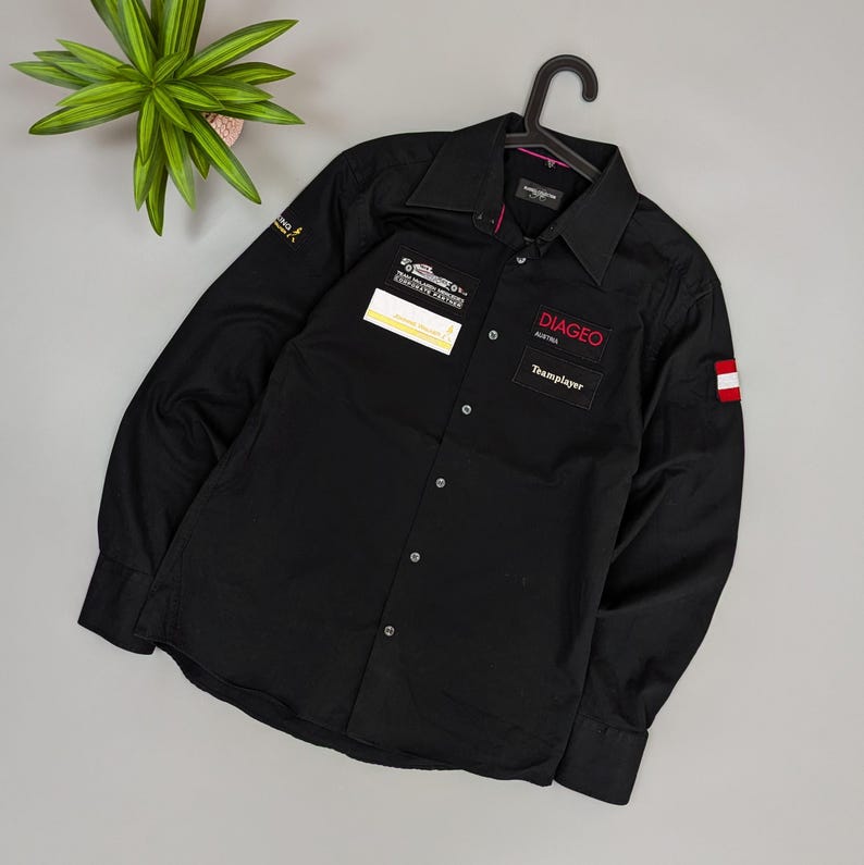 Puede incluir: Camisa negra de manga larga con cuello. Presenta parches con texto y logotipos, incluyendo "DIAGEO AUSTRIA Teamplayer" en rojo. Un parche con la bandera austriaca est&aacute; en la manga derecha. La camisa est&aacute; colgada de una percha negra.