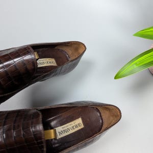 Może przedstawiać: Para brązowych sk&oacute;rzanych mokasyn&oacute;w o fakturze sk&oacute;ry krokodyla. Buty mają klasyczny design z zaokrąglonym noskiem i niskim obcasem. Logo Giorgio Armani jest widoczne wewnątrz buta.