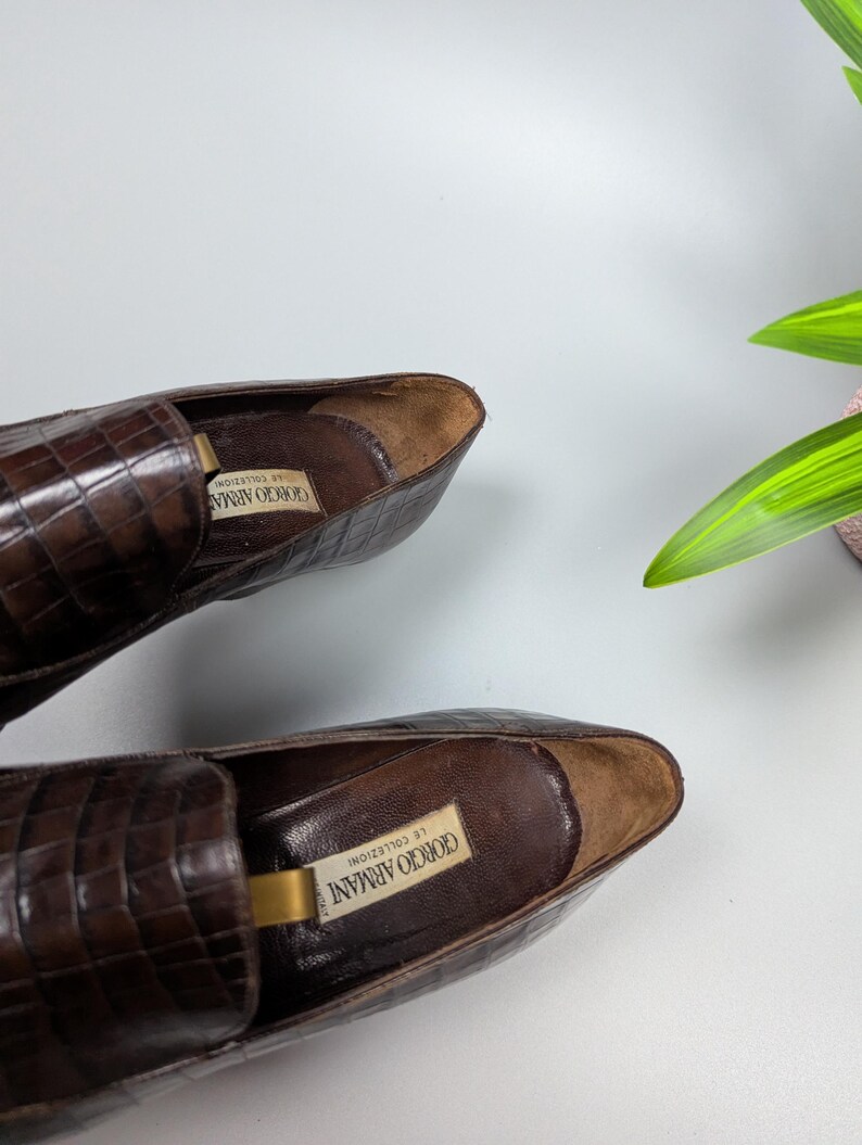 Może przedstawiać: Para brązowych sk&oacute;rzanych mokasyn&oacute;w o fakturze sk&oacute;ry krokodyla. Buty mają klasyczny design z zaokrąglonym noskiem i niskim obcasem. Logo Giorgio Armani jest widoczne wewnątrz buta.