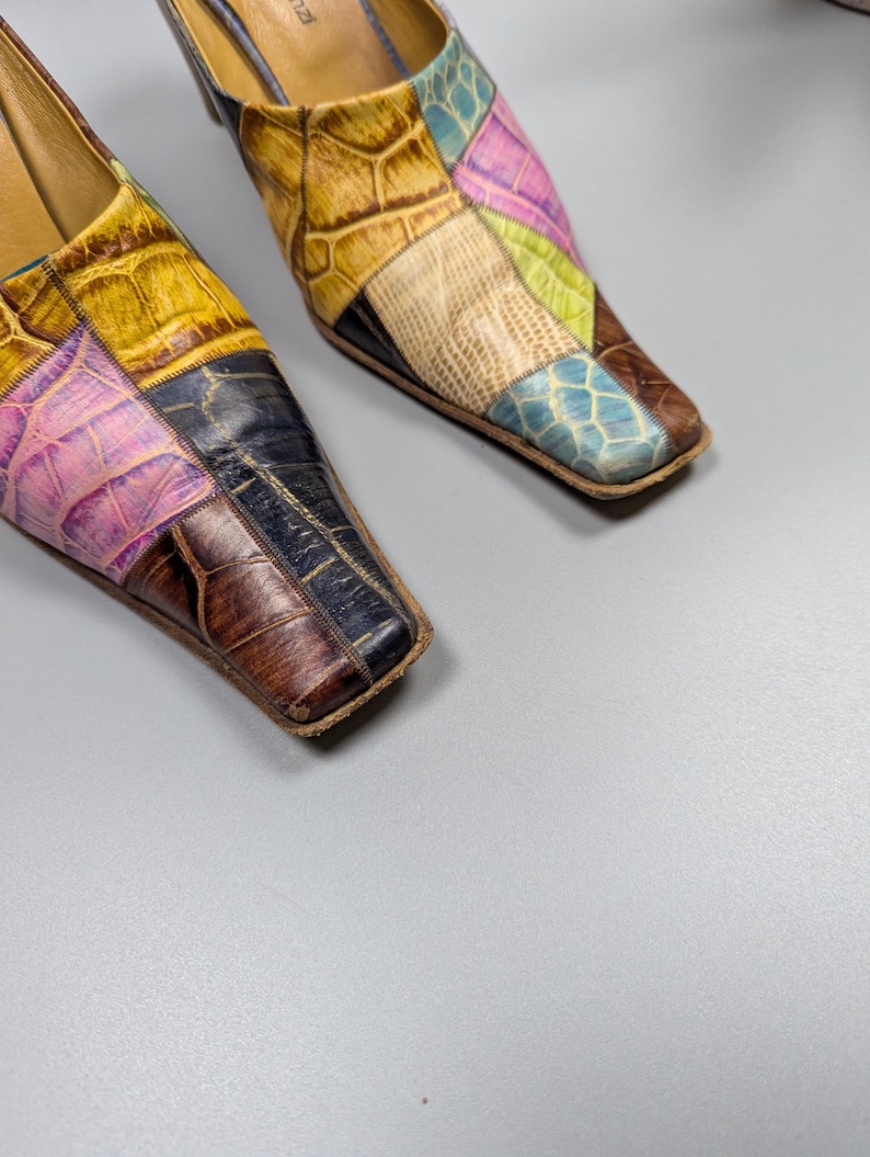 May include: A pair of colorful patchwork leather mules. The shoes feature a square toe and a low heel. The patchwork design includes shades of yellow, pink, blue, and brown, creating a unique and eye-catching pattern.