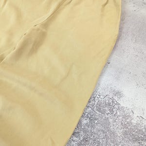 May include: A pair of light yellow trousers featuring a smooth, solid-colored fabric and an elasticized waistband. The garment is constructed with a simple, clean design, showcasing a consistent light yellow hue throughout the visible portion of the pants.