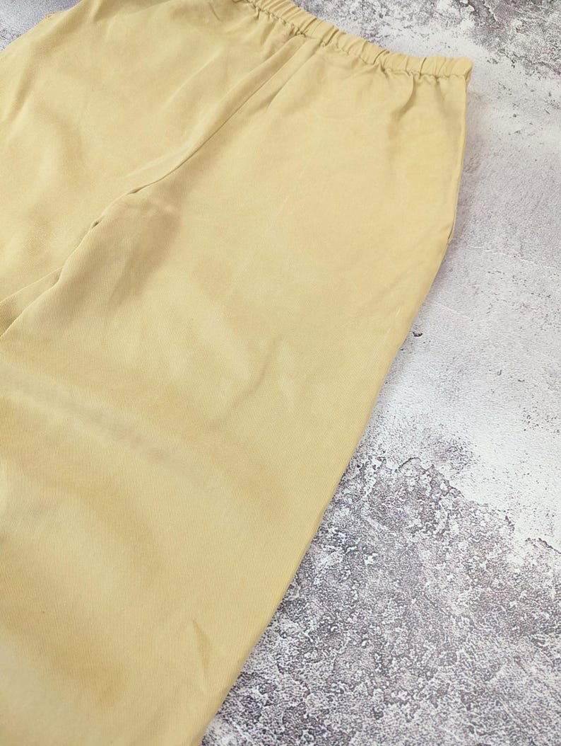 May include: A pair of light yellow trousers featuring a smooth, solid-colored fabric and an elasticized waistband. The garment is constructed with a simple, clean design, showcasing a consistent light yellow hue throughout the visible portion of the pants.