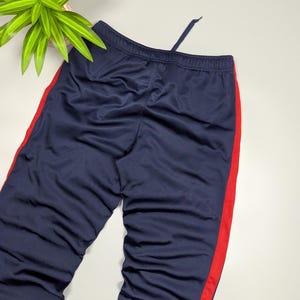 May include: Navy blue tracksuit bottoms with a red stripe down the side. The trousers have a drawstring at the waist. The fabric appears to be a smooth, synthetic material.