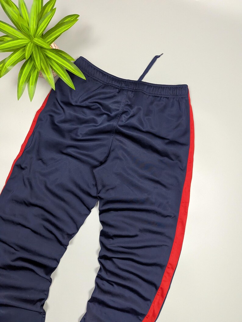 May include: Navy blue tracksuit bottoms with a red stripe down the side. The trousers have a drawstring at the waist. The fabric appears to be a smooth, synthetic material.