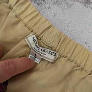 May include: A pair of tan trousers with an elastic waistband. The interior label reads Michael Glasser for Democracy U.S.A. and includes a size L tag with the text Made in U.S.A. The fabric has a visible diagonal twill weave texture throughout.