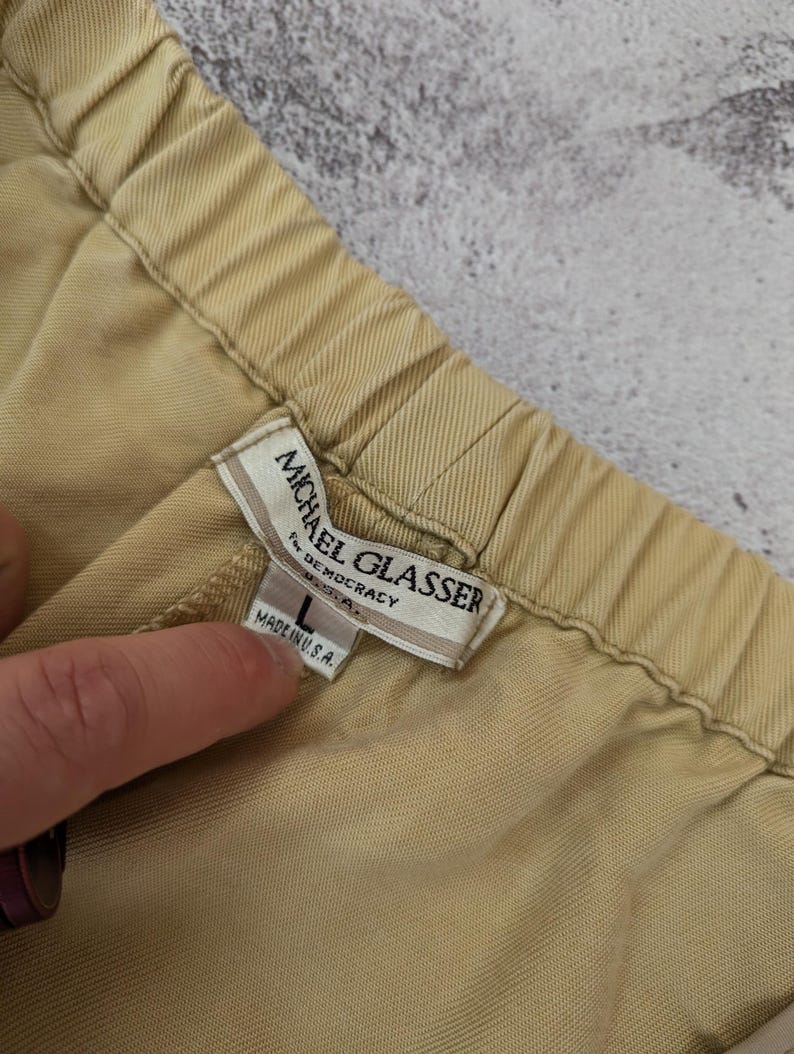 May include: A pair of tan trousers with an elastic waistband. The interior label reads Michael Glasser for Democracy U.S.A. and includes a size L tag with the text Made in U.S.A. The fabric has a visible diagonal twill weave texture throughout.