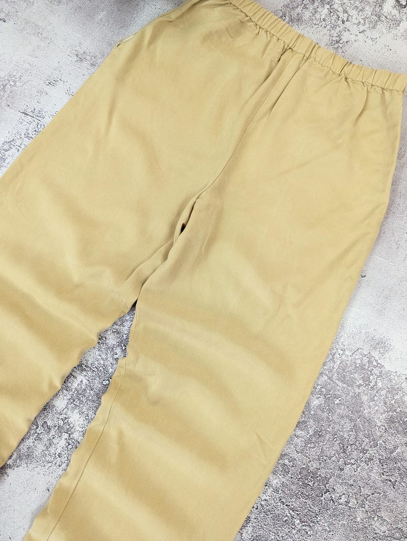 May include: A pair of solid light tan trousers featuring a comfortable elasticized waistband. The pants are crafted from a smooth, plain-weave fabric and showcase a simple, straight-leg cut with clean lines and no visible pockets or decorative details.