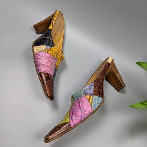 May include: A pair of patchwork leather mules with a pointed toe and wooden block heel. The shoes feature a mix of colors including brown, pink, yellow, blue, and black. The heels are approximately 3 inches high.