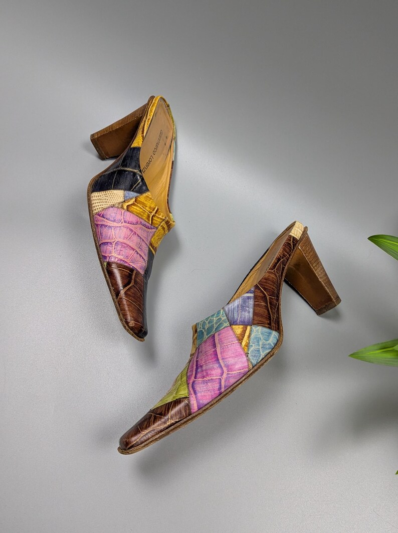 May include: A pair of patchwork leather mules with a pointed toe and wooden block heel. The shoes feature a mix of colors including brown, pink, yellow, blue, and black. The heels are approximately 3 inches high.