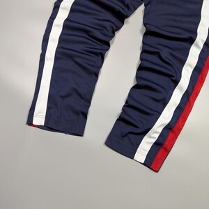 May include: Navy blue track pants with white side stripes and a small Nike logo. The pants have a red accent stripe on one leg. The trousers are made of a smooth, possibly synthetic material.