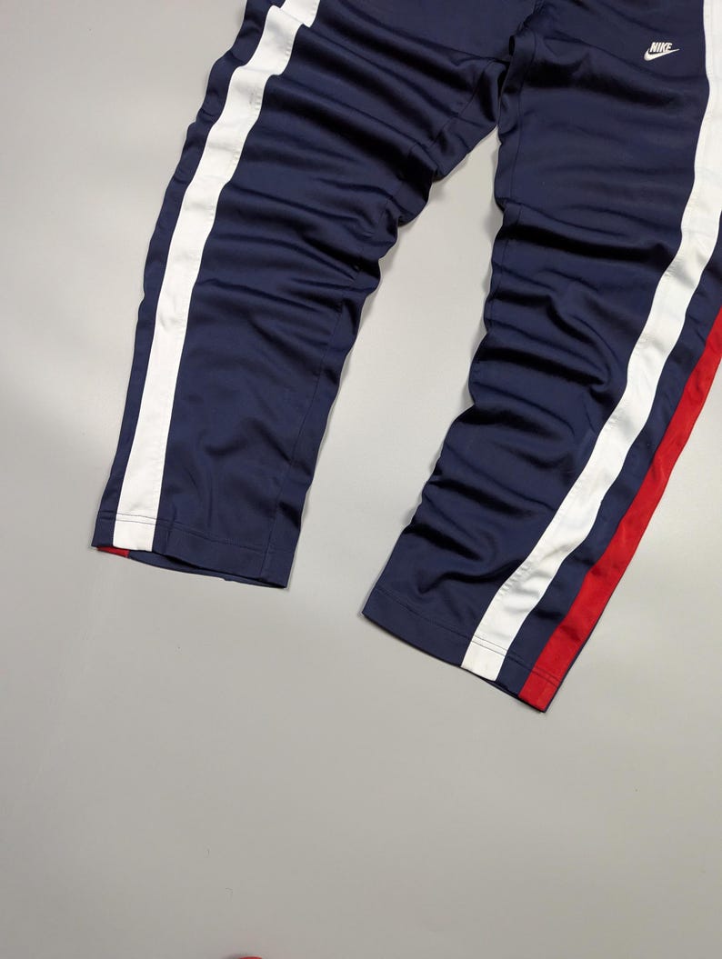 May include: Navy blue track pants with white side stripes and a small Nike logo. The pants have a red accent stripe on one leg. The trousers are made of a smooth, possibly synthetic material.