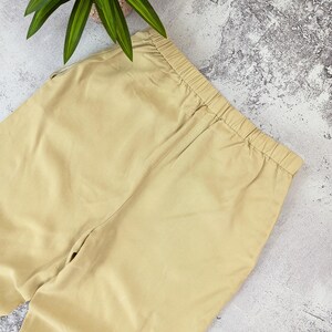 May include: A pair of solid beige trousers featuring an elasticized waistband with gathered fabric detailing. The pants are constructed from a smooth, plain-weave material and display a consistent light tan color throughout the visible garment area.