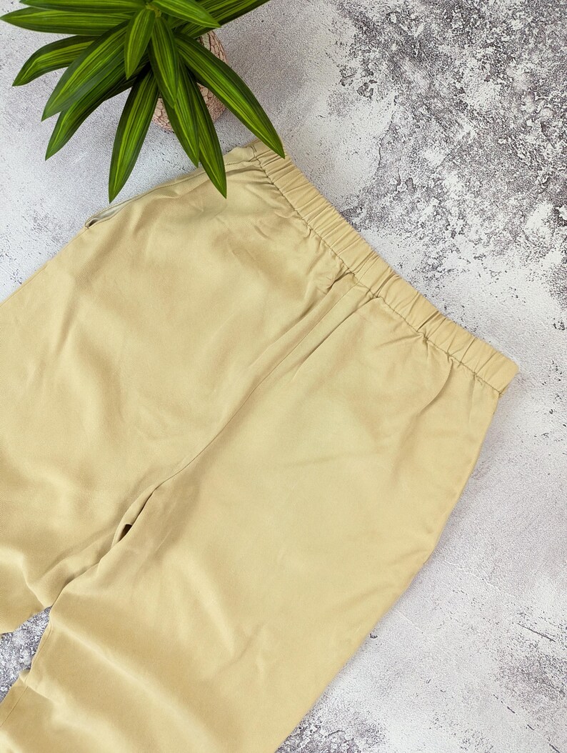 May include: A pair of solid beige trousers featuring an elasticized waistband with gathered fabric detailing. The pants are constructed from a smooth, plain-weave material and display a consistent light tan color throughout the visible garment area.