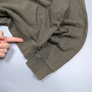 Puede incluir: Primer plano de una sudadera con capucha verde oliva descolorida. La sudadera tiene un bolsillo de canguro y pu&ntilde;os y dobladillo acanalados. La tela parece desgastada y tiene una est&eacute;tica vintage. La imagen est&aacute; tomada sobre un fondo gris claro.