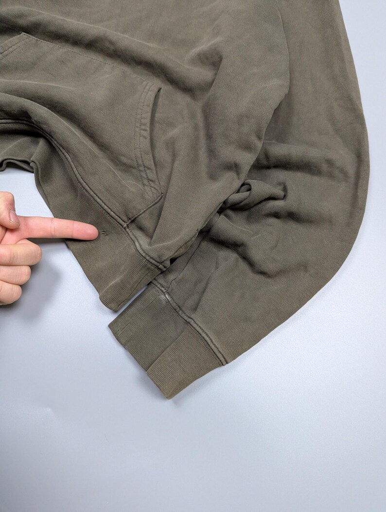 Puede incluir: Primer plano de una sudadera con capucha verde oliva descolorida. La sudadera tiene un bolsillo de canguro y pu&ntilde;os y dobladillo acanalados. La tela parece desgastada y tiene una est&eacute;tica vintage. La imagen est&aacute; tomada sobre un fondo gris claro.