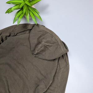 Puede incluir: Una sudadera con capucha verde oliva con capucha con cord&oacute;n. La sudadera est&aacute; hecha de una tela suave y texturizada. Una planta verde y amarilla est&aacute; en el fondo.