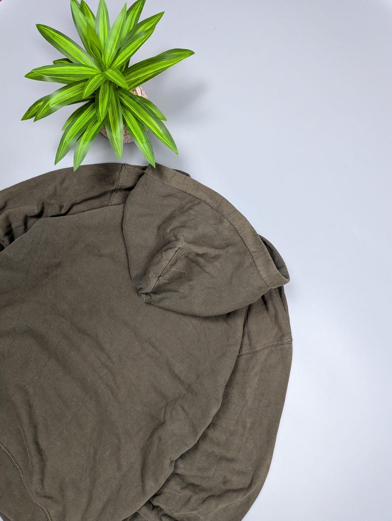 Puede incluir: Una sudadera con capucha verde oliva con capucha con cord&oacute;n. La sudadera est&aacute; hecha de una tela suave y texturizada. Una planta verde y amarilla est&aacute; en el fondo.