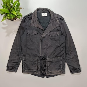 Puede incluir: Una chaqueta de algodón negra con cierre de botones, múltiples bolsillos y cuello abatible. La chaqueta tiene una etiqueta que dice "L.A.S.S.I.C."