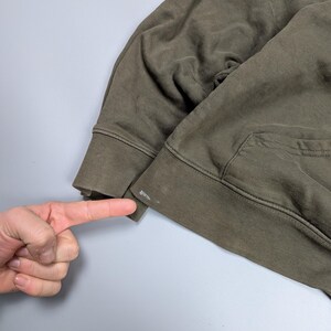 Puede incluir: Sudadera con capucha verde oliva con bolsillo de canguro y pu&ntilde;os acanalados. La imagen muestra un primer plano de la manga y el pu&ntilde;o de la sudadera con capucha. La tela parece ser un material suave y desgastado.