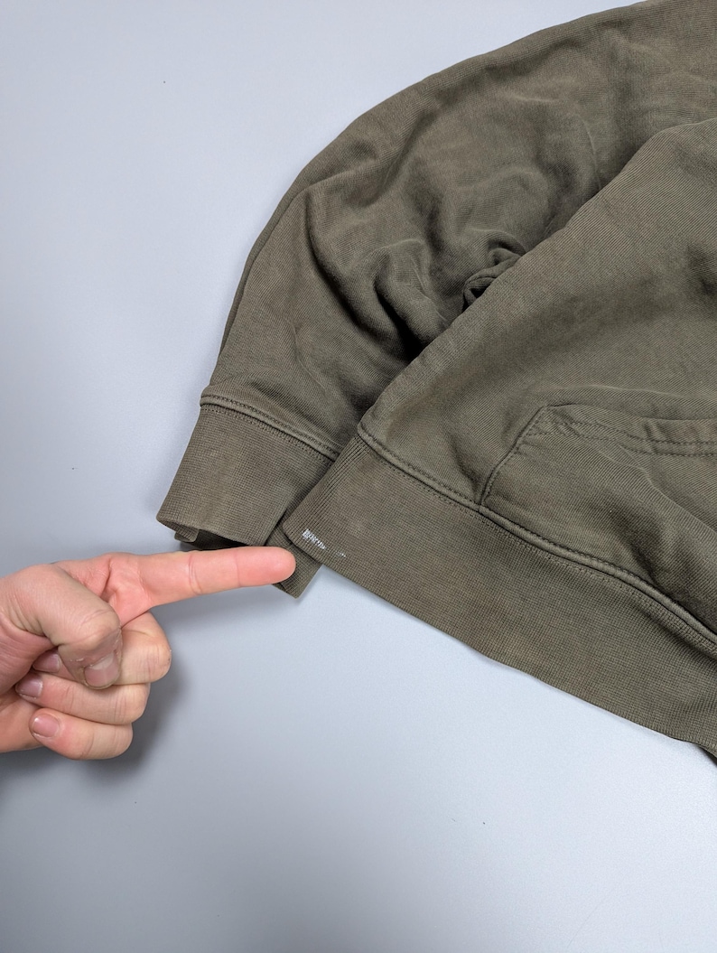 Puede incluir: Sudadera con capucha verde oliva con bolsillo de canguro y pu&ntilde;os acanalados. La imagen muestra un primer plano de la manga y el pu&ntilde;o de la sudadera con capucha. La tela parece ser un material suave y desgastado.