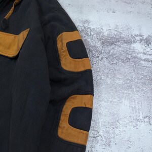Pode incluir: Um casaco preto com capuz apresentando remendos em tom castanho nos ombros e cotovelos. A pe&ccedil;a inclui um fecho de correr frontal, um capuz com cord&atilde;o e acabamento castanho, e um design estruturado com pain&eacute;is de tecido refor&ccedil;ado na manga.