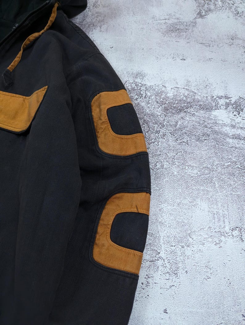 Pode incluir: Um casaco preto com capuz apresentando remendos em tom castanho nos ombros e cotovelos. A pe&ccedil;a inclui um fecho de correr frontal, um capuz com cord&atilde;o e acabamento castanho, e um design estruturado com pain&eacute;is de tecido refor&ccedil;ado na manga.