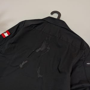 Puede incluir: Camisa negra abotonada con una figura bordada en la espalda. Un peque&ntilde;o parche de la bandera austriaca es visible en la manga izquierda. La camisa est&aacute; colgada de una percha negra.