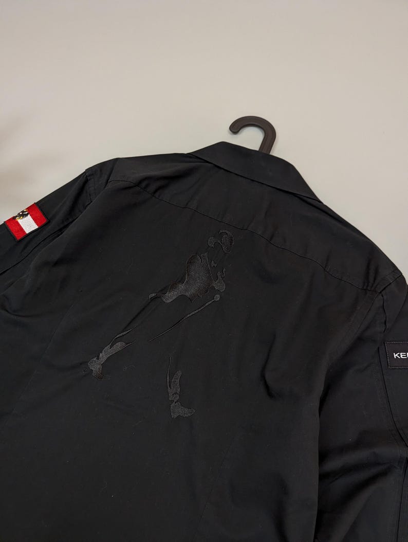 Puede incluir: Camisa negra abotonada con una figura bordada en la espalda. Un peque&ntilde;o parche de la bandera austriaca es visible en la manga izquierda. La camisa est&aacute; colgada de una percha negra.
